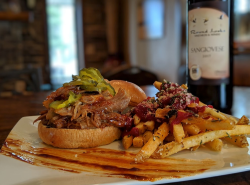 A pulled pork sandwich with pickles, served with seasoned fries and a bottle of Sangiovese wine in the background.