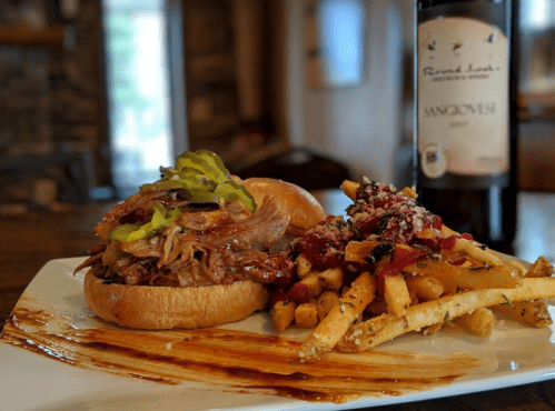 A pulled pork sandwich with pickles, served with seasoned fries and a bottle of Sangiovese wine in the background.