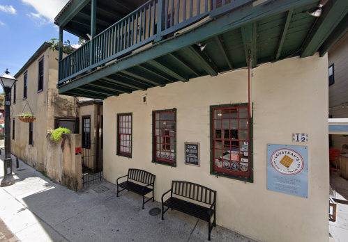 A quaint building with green trim, featuring two benches and a sign, set in a sunny street with a balcony above.