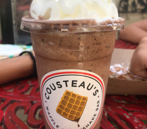 A chocolate milkshake topped with whipped cream in a clear cup, featuring a waffle cone logo on the label.