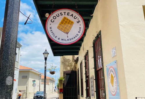 Sign for Cousteau's Waffle & Milkshake Bar hanging from a building, with a street view in the background.