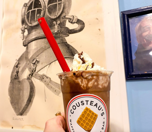 A hand holding a chocolate milkshake topped with whipped cream, with a vintage diving suit illustration in the background.