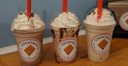 Three milkshakes in clear cups with whipped cream and colorful straws, labeled "Cousteau's Waffles & Milkshakes."