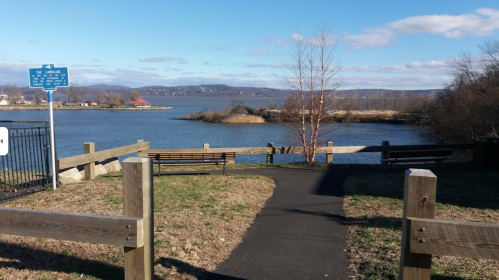 A scenic view of a river with benches and a pathway, surrounded by trees and distant hills under a clear blue sky.