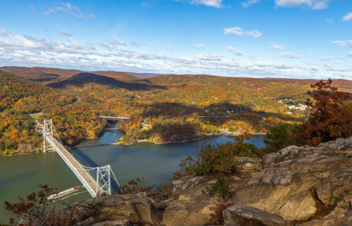 A scenic view of a bridge over a river, surrounded by colorful autumn foliage and rolling hills under a blue sky.