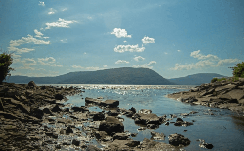 A serene river scene with rocky shores, mountains in the background, and a bright blue sky with scattered clouds.