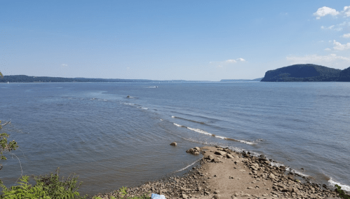 A serene river scene with gentle waves, rocky shore, and distant hills under a clear blue sky.