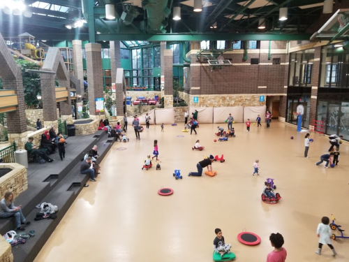 A spacious indoor play area filled with children playing on toys, with adults seated nearby. Bright and lively atmosphere.