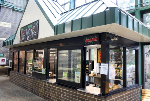 A modern food kiosk with a green roof and large windows, displaying a menu and food items inside.