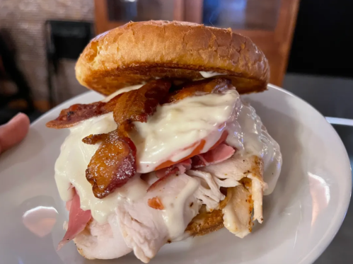 A close-up of a hearty sandwich with turkey, ham, bacon, and creamy sauce on a toasted bun, served on a plate.