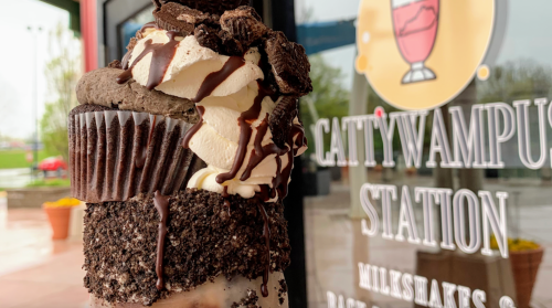 A towering dessert featuring a cupcake, whipped cream, crushed Oreos, and chocolate drizzle in front of a shop window.