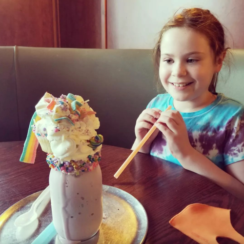 A smiling child sits at a table, excitedly looking at a colorful milkshake topped with whipped cream and sprinkles.