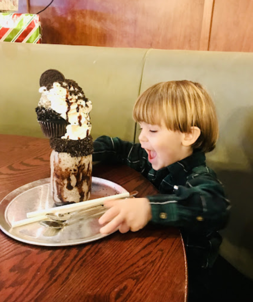A joyful child with a big milkshake topped with a cupcake, sitting at a table with a silver tray and chopsticks.