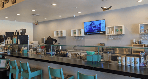 A cozy café interior with a counter, bar stools, and a TV displaying a show. A staff member is preparing drinks.