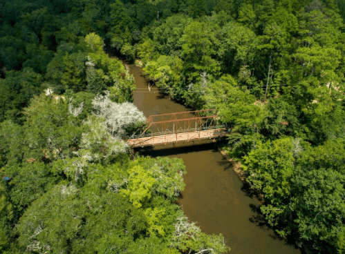 Aerial view of a rusted bridge over a winding river, surrounded by dense green trees and foliage.