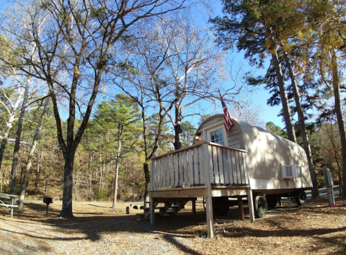 A small trailer with a porch, surrounded by trees in a wooded area, under a clear blue sky.
