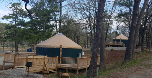 Two yurts with tan roofs on wooden decks, surrounded by trees in a serene outdoor setting.