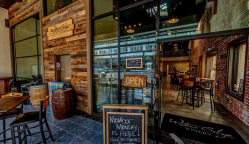 Exterior view of a winery with a welcoming "Open" sign and rustic decor, featuring large glass doors and wooden accents.