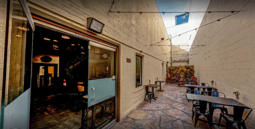 A narrow outdoor patio with stone flooring, tables, and string lights, flanked by brick walls and a colorful mural.