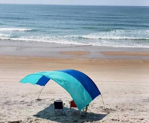 A beach scene featuring a blue and teal canopy over two chairs, with gentle waves and a sandy shore in the background.