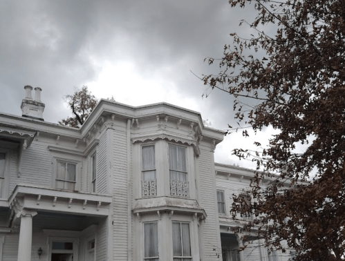 A historic white house with ornate details under a cloudy sky, surrounded by bare trees.