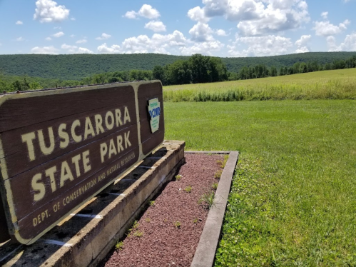 Sign for Tuscarora State Park with green fields and hills in the background under a blue sky with fluffy clouds.