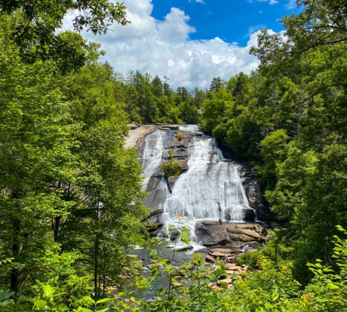 A scenic waterfall cascades down rocky cliffs, surrounded by lush green trees and a bright blue sky.