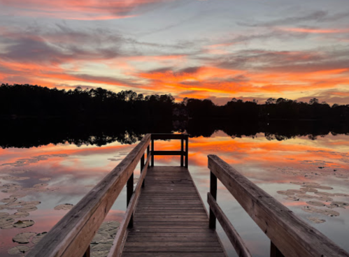 A wooden pier extends into a calm lake at sunset, reflecting vibrant orange and pink hues in the water.