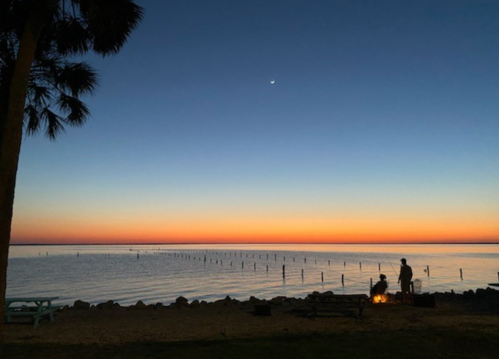 Silhouette of a person by a fire on the beach at sunset, with a calm sea and a crescent moon in the sky.