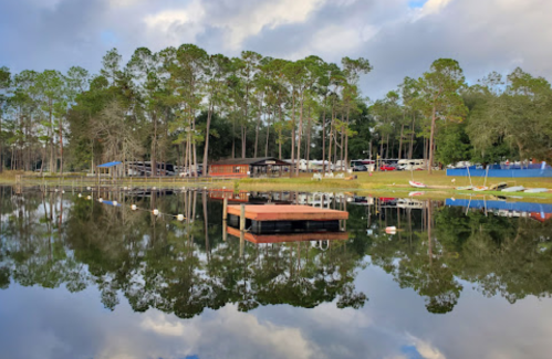 A serene lake scene with a wooden dock, surrounded by trees and reflections of the sky and buildings on the water.