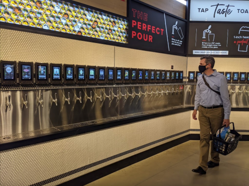 A man in a mask walks past a wall of digital taps and screens in a modern beverage dispensing area.
