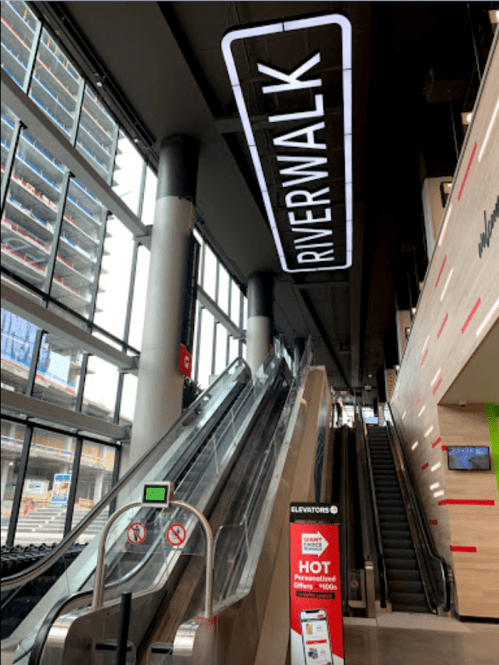 Interior view of a modern building featuring an escalator, a "RIVERWALK" sign, and an elevator information display.