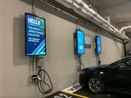 Three digital screens in a parking garage display information about free electric vehicle charging. A car is plugged in.