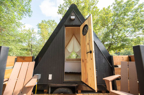 A black A-frame cabin with an open door, surrounded by trees and wooden chairs on a deck.