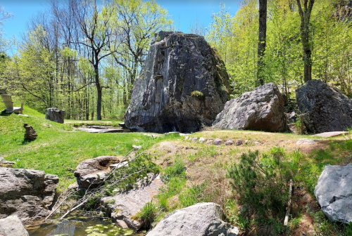A serene landscape featuring a large rock formation surrounded by greenery and trees under a clear blue sky.