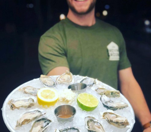 A person holds a platter of oysters on ice, garnished with lemon, lime, and dipping sauces.
