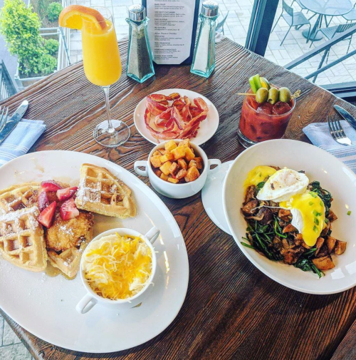 A table set with waffles topped with strawberries, a savory dish with eggs and greens, potatoes, and drinks.