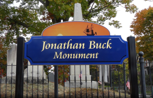 Sign for the Jonathan Buck Monument, featuring a ship silhouette and surrounded by trees and gravestones.