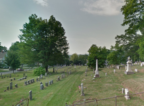 A peaceful cemetery with gravestones and large trees under a cloudy sky.