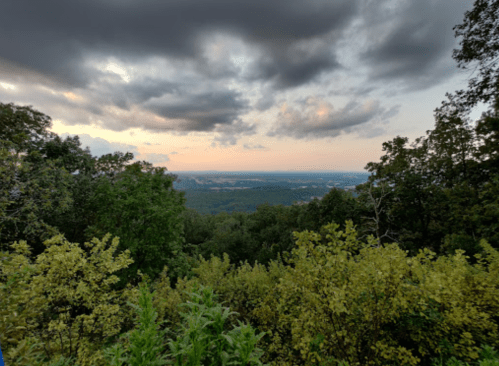 A scenic view of a valley at sunset, surrounded by lush green trees and a cloudy sky.