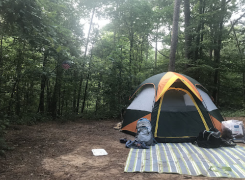 A colorful tent set up in a wooded campsite, with a backpack and a mat on the ground nearby.