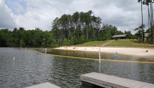 A serene lake scene with a sandy beach, people relaxing, and trees in the background under a cloudy sky.