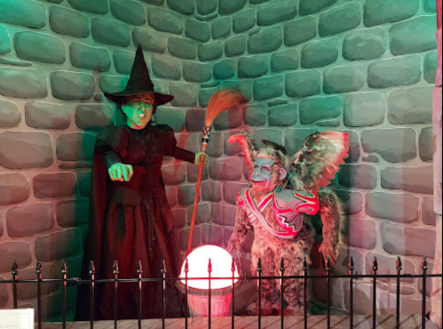 A witch in a black dress and hat stands next to a winged creature, with a glowing orb between them, against a stone wall.