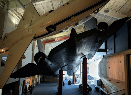 A large aircraft and a black jet hang from the ceiling of a spacious museum exhibit. Visitors are seen below.