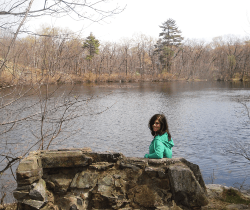 A woman in a green jacket sits on a stone wall by a calm lake surrounded by bare trees.