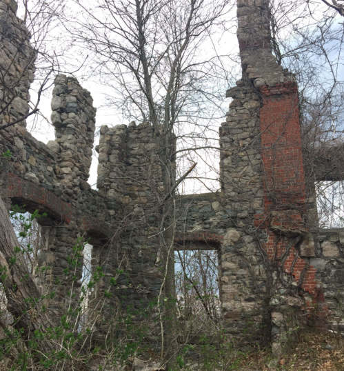 Ruins of a stone and brick structure, overgrown with trees and vegetation, against a cloudy sky.