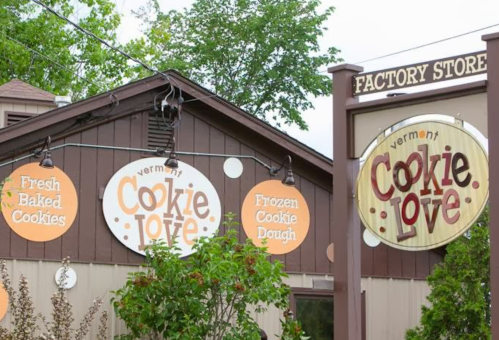 Signage for "Vermont Cookie Love" factory store, featuring fresh baked cookies and frozen cookie dough. Green trees in background.