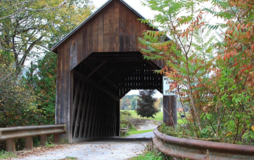 A wooden covered bridge surrounded by trees and autumn foliage, leading to a rural path.