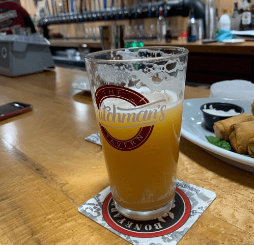 A glass of hazy beer sits on a wooden bar top, with a logo and a coaster underneath. In the background, taps are visible.