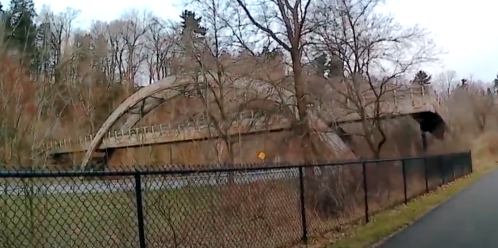 A wooden arch bridge surrounded by trees, with a path and fence in the foreground.